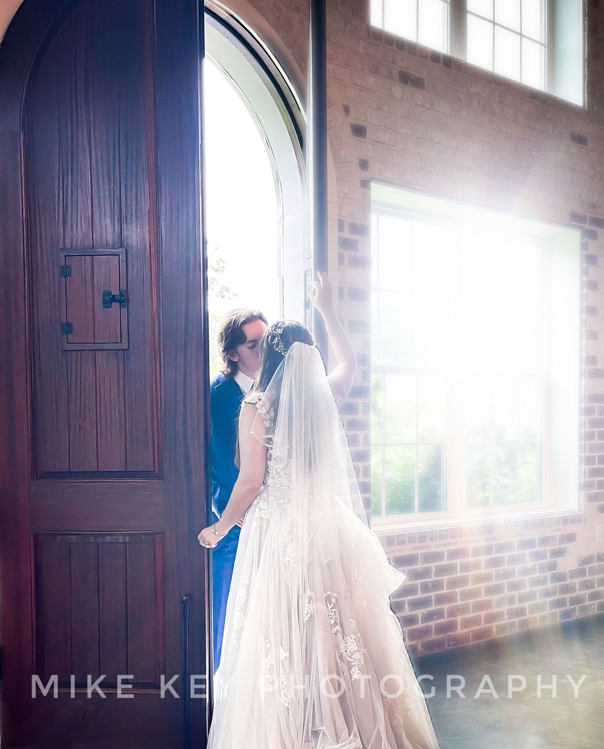 A bride in a white wedding gown and a person in a blue suit stand together in a sunlit, arched doorway.