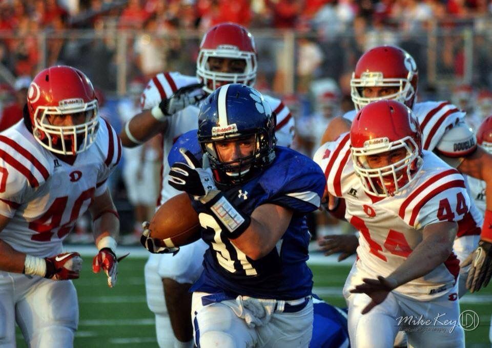 A football player in a blue jersey runs with the ball, pursued by opponents in white jerseys on a green field.