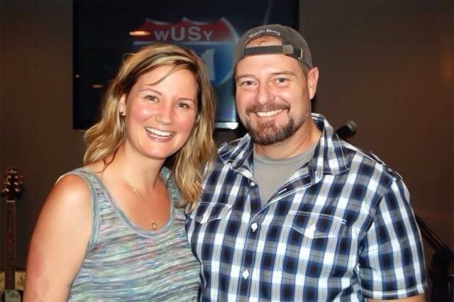 A smiling woman and a man in a plaid shirt and cap pose together in a radio studio with a station logo in the background.