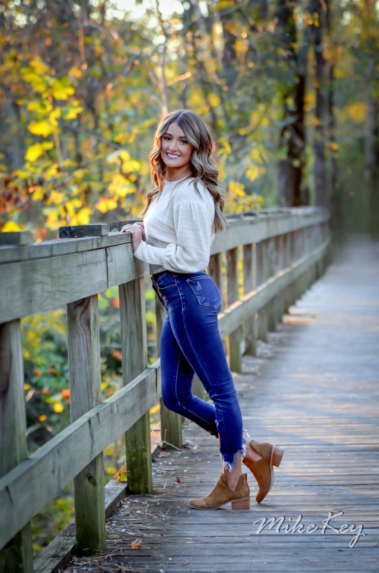 A person smiling while leaning against a wooden bridge railing in a sunlit, tree-lined forest setting.