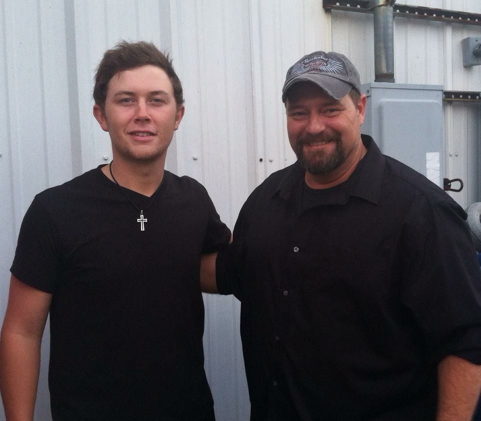 Two individuals standing side-by-side, smiling, in front of a white corrugated metal wall.
