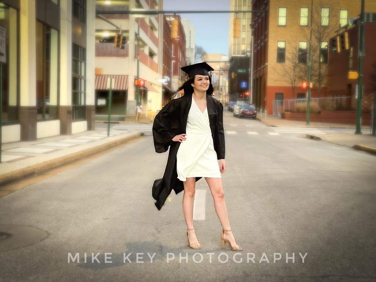 A graduate in a cap, gown, and white dress poses on a city street.