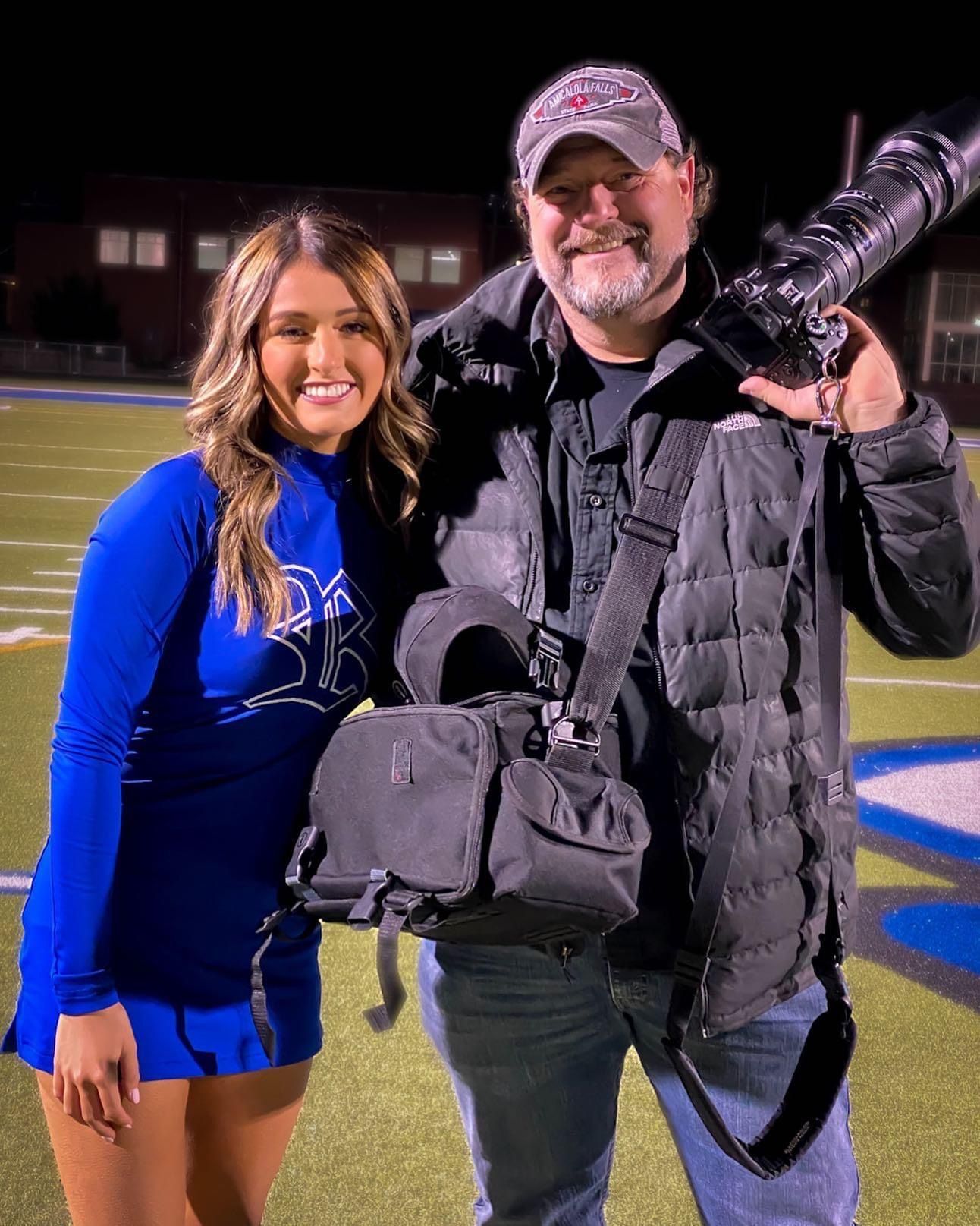 A cheerleader in a blue uniform and a photographer with camera gear smile together on a football field at night.