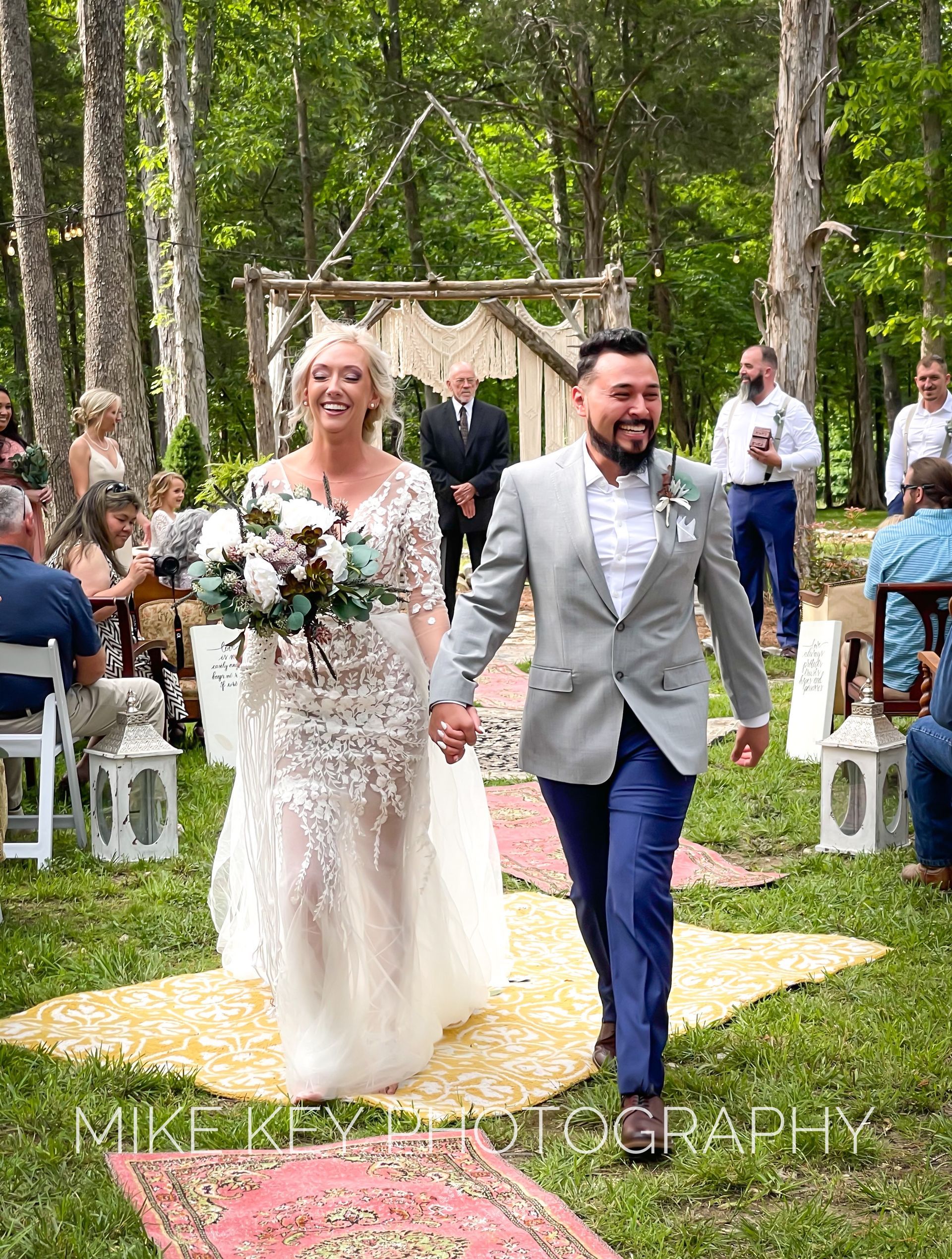 A smiling couple holding hands walks down a fabric aisle in a wooded setting after a wedding ceremony.