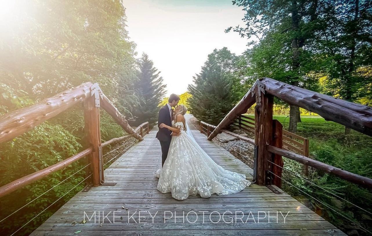 A couple in wedding attire embraces on a rustic wooden bridge amidst a sunlit, tree-lined forest setting.