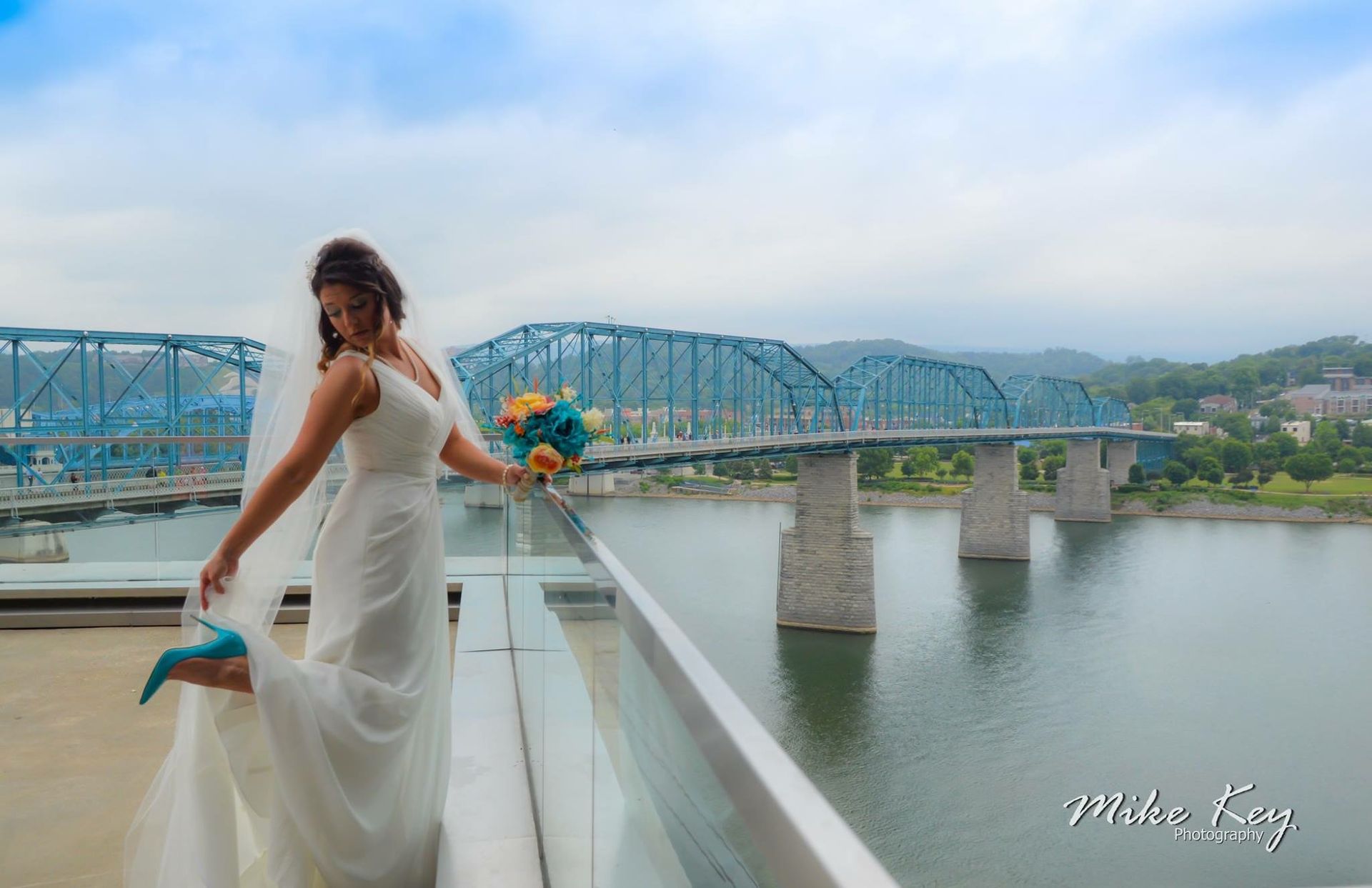 A bride in a white wedding gown holding a bouquet stands on a terrace, showing her teal heels with a blue bridge behind.