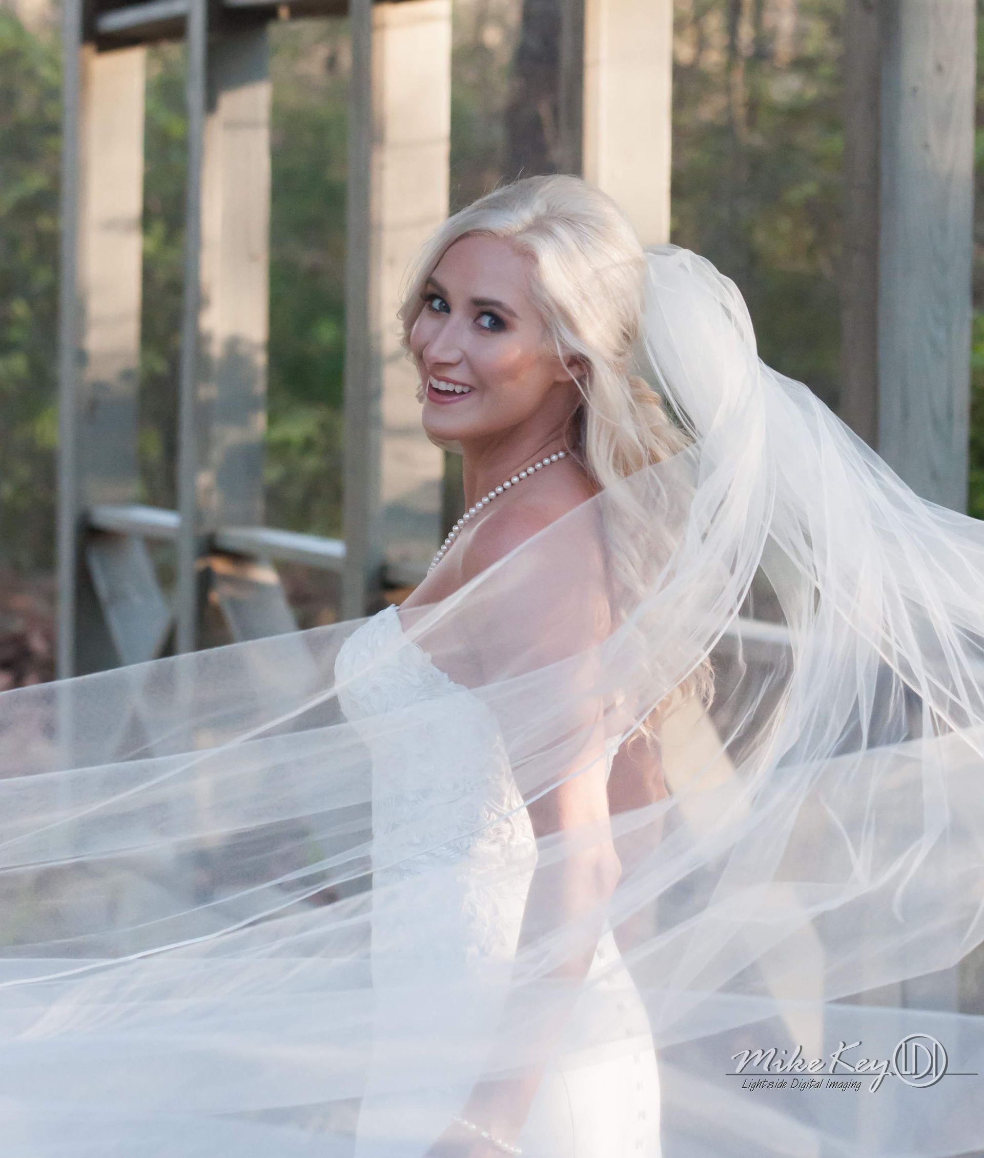 A bride in a white wedding dress and veil smiling while looking over her shoulder against an outdoor rustic backdrop.