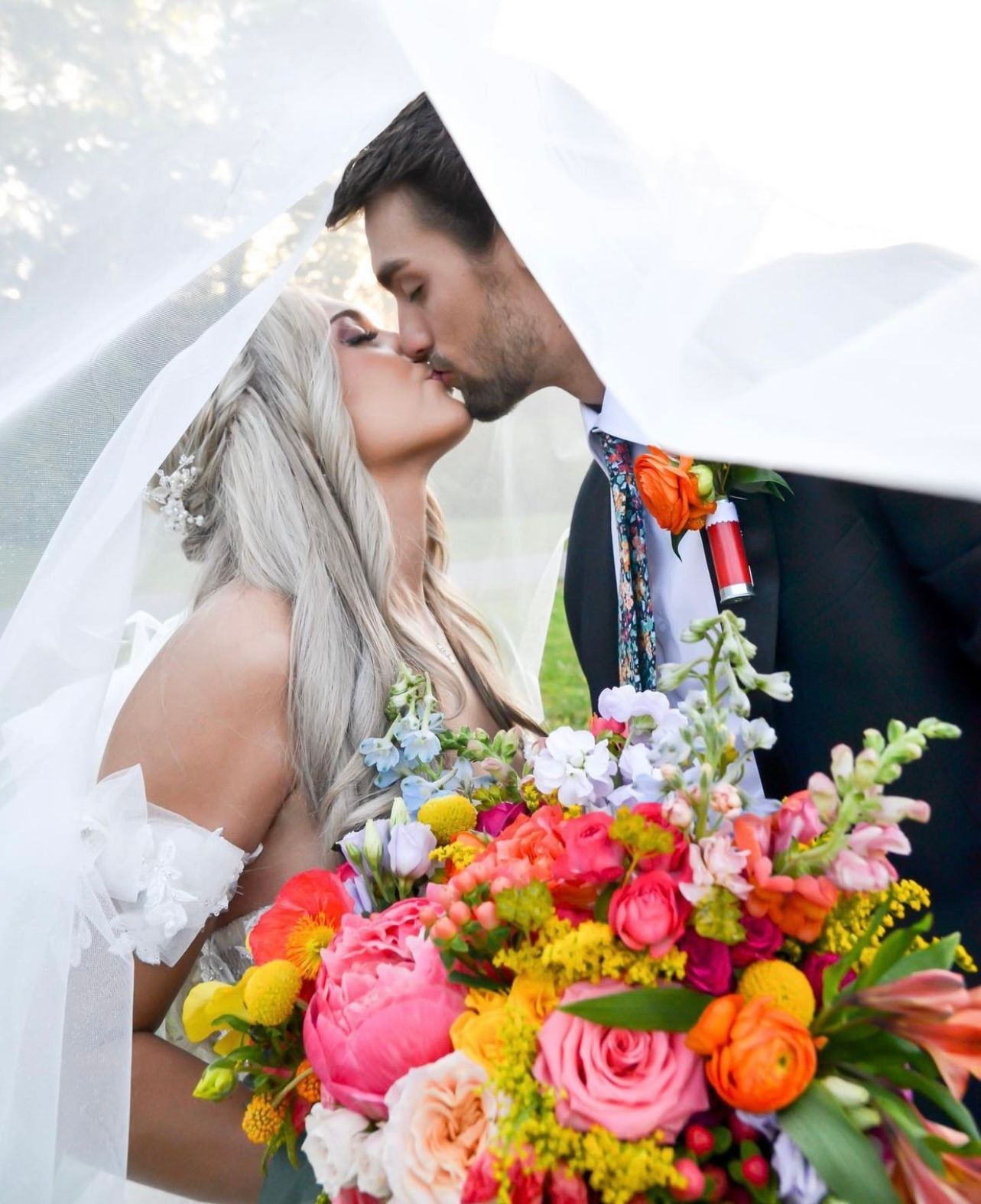 A bride and groom kiss under a wedding veil, holding a large, colorful bouquet of bright flowers outdoors.