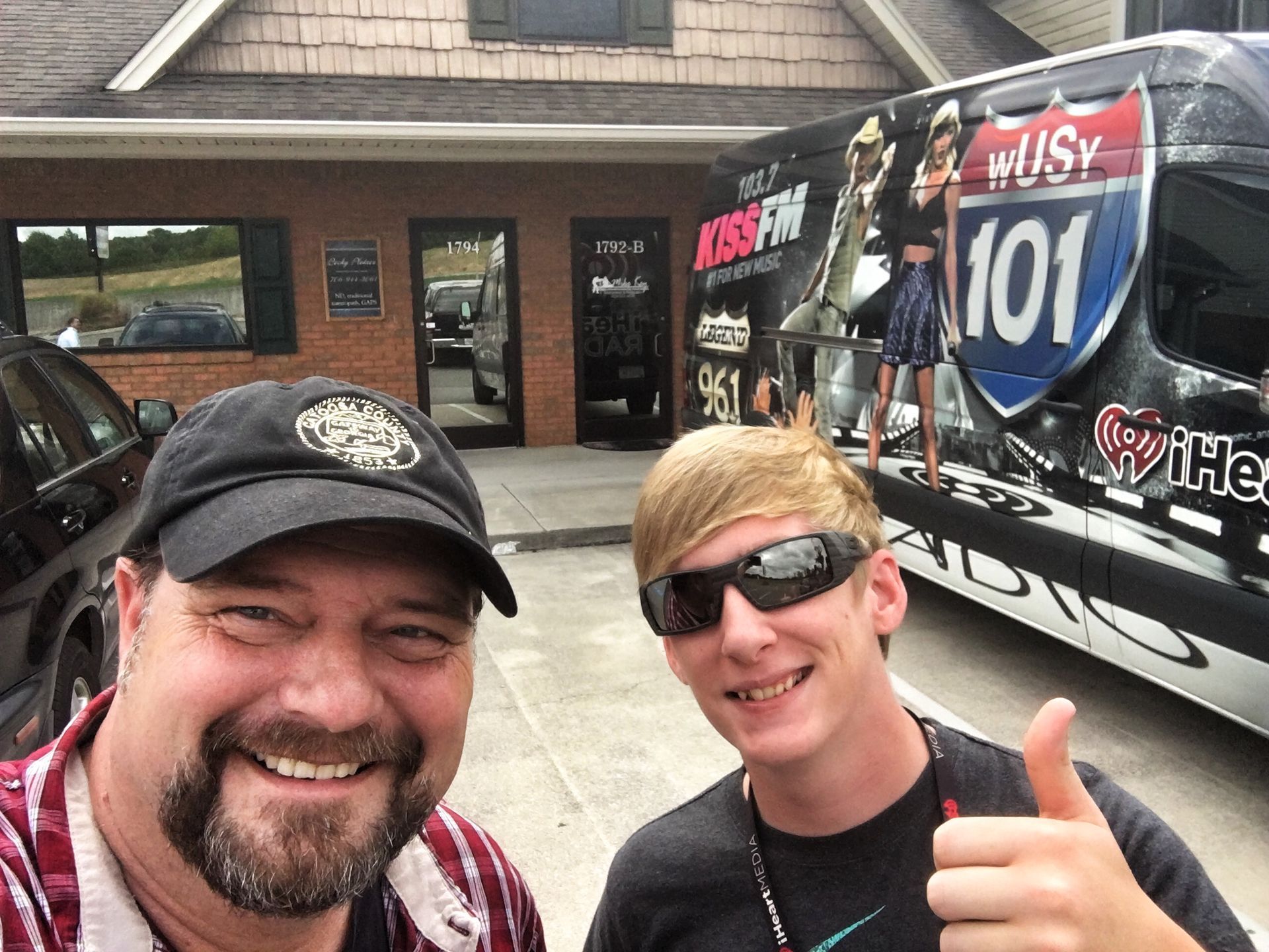 Two people smile for a selfie in front of a brick building and a radio station branded van with a 101 logo.