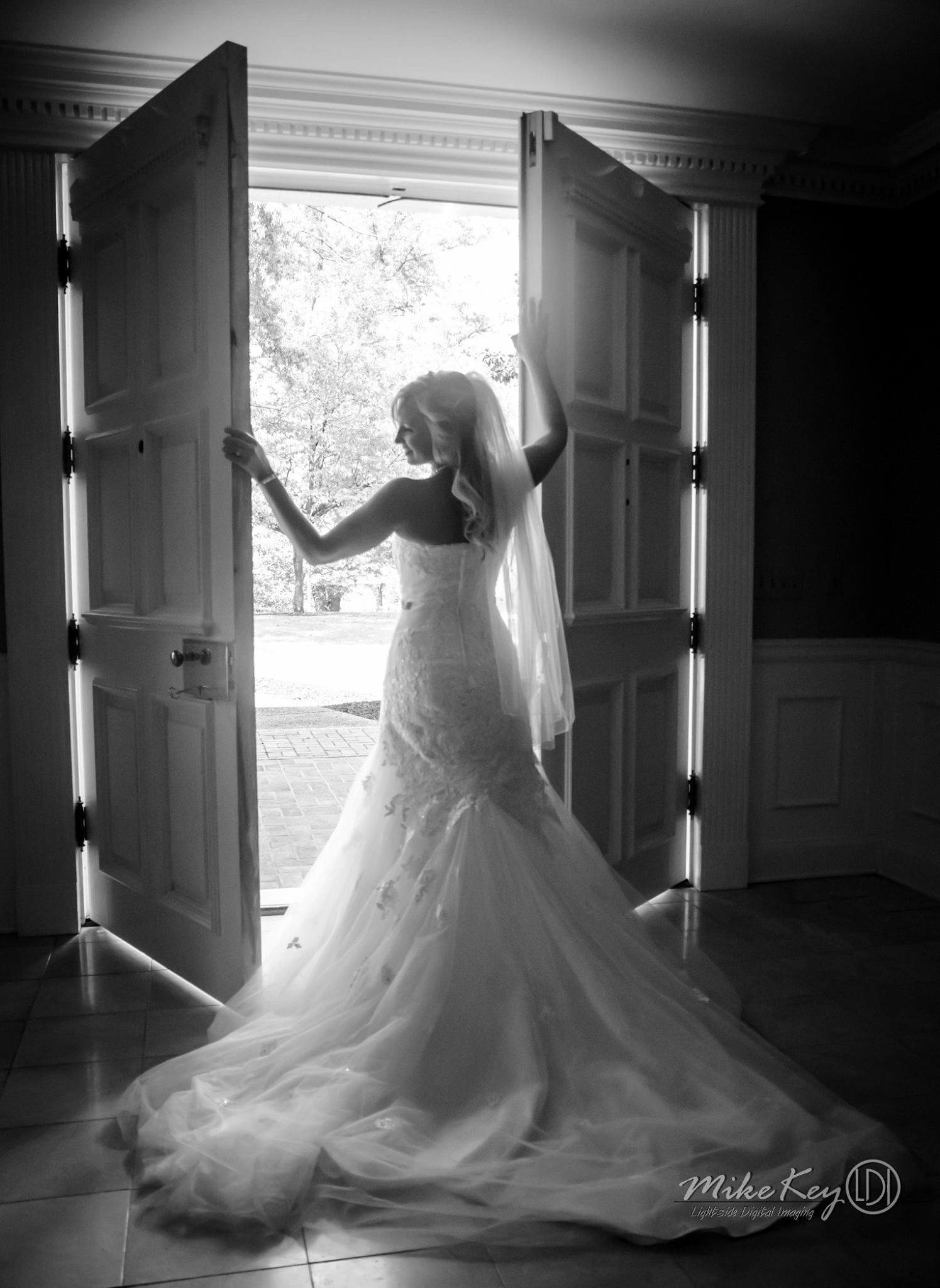 A bride in a long, white wedding dress stands in a doorway, silhouetted against bright light.