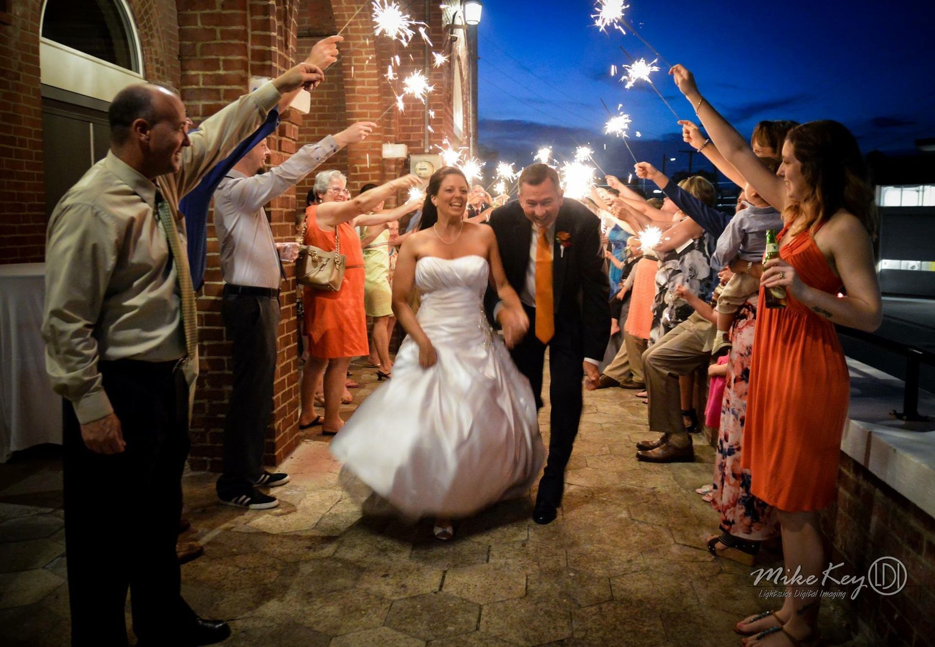 A bride and groom walk through a line of guests holding lit sparklers outside a brick building at twilight.