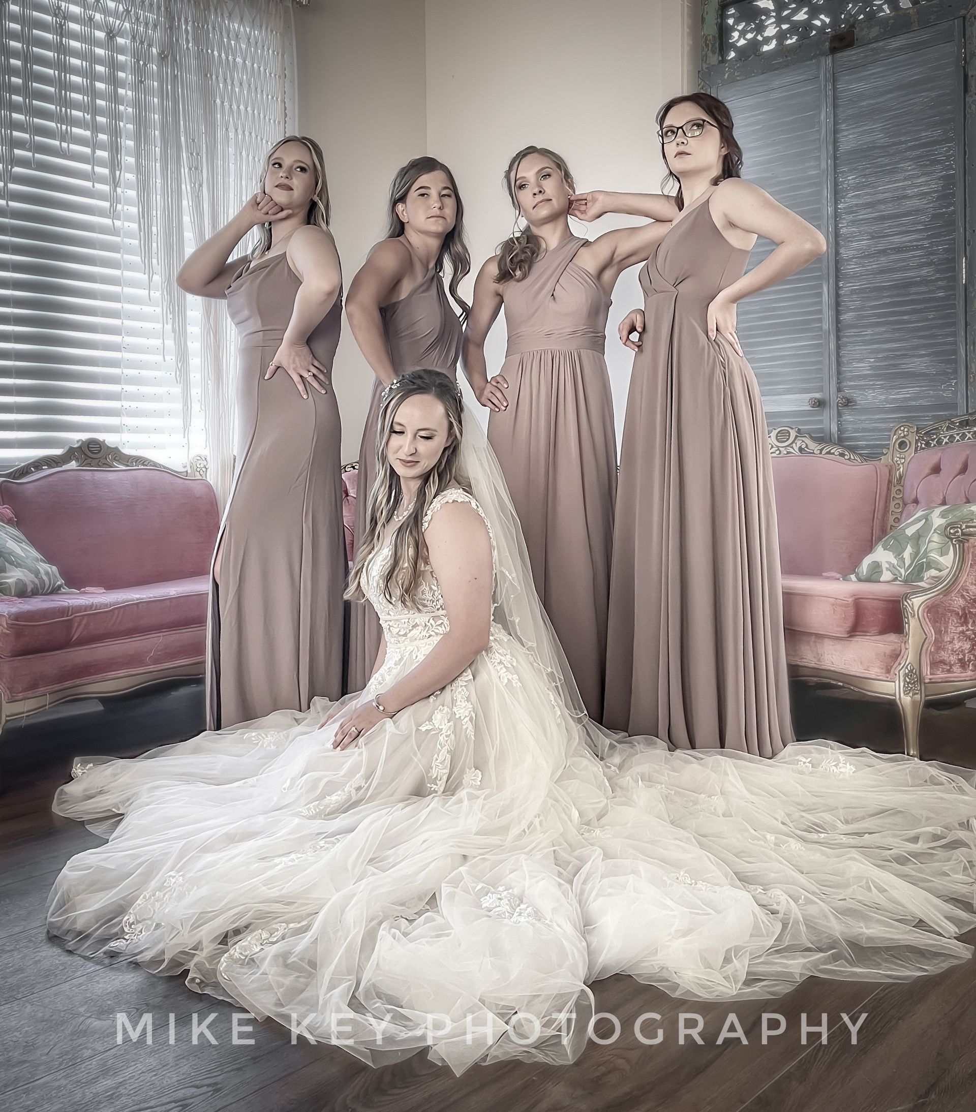 A bride in a white lace gown sits on the floor before four bridesmaids in floor-length, mauve dresses in a room.