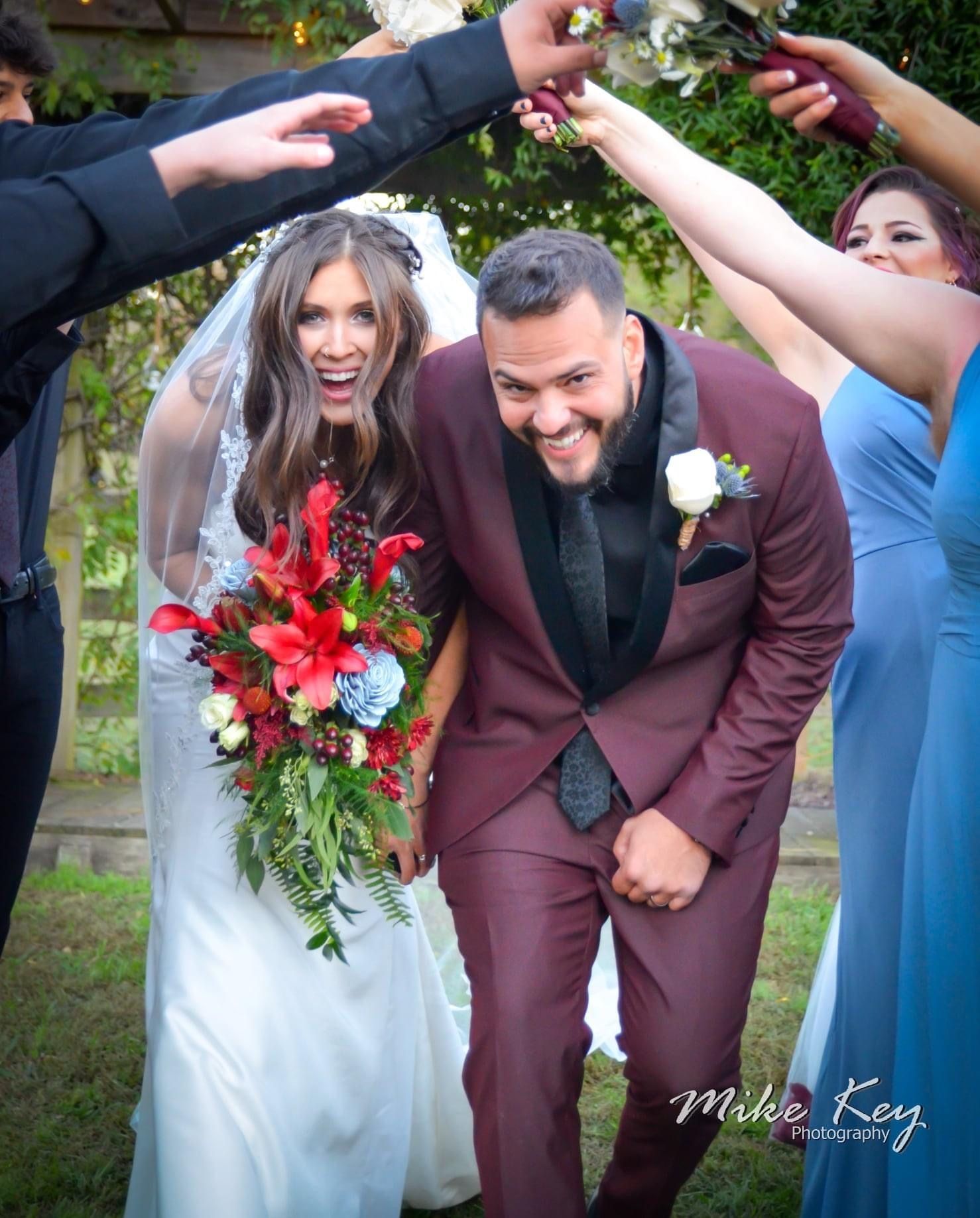 A smiling bride and groom in a suit exit a tunnel of people's arms, holding a vibrant bouquet, outdoors.