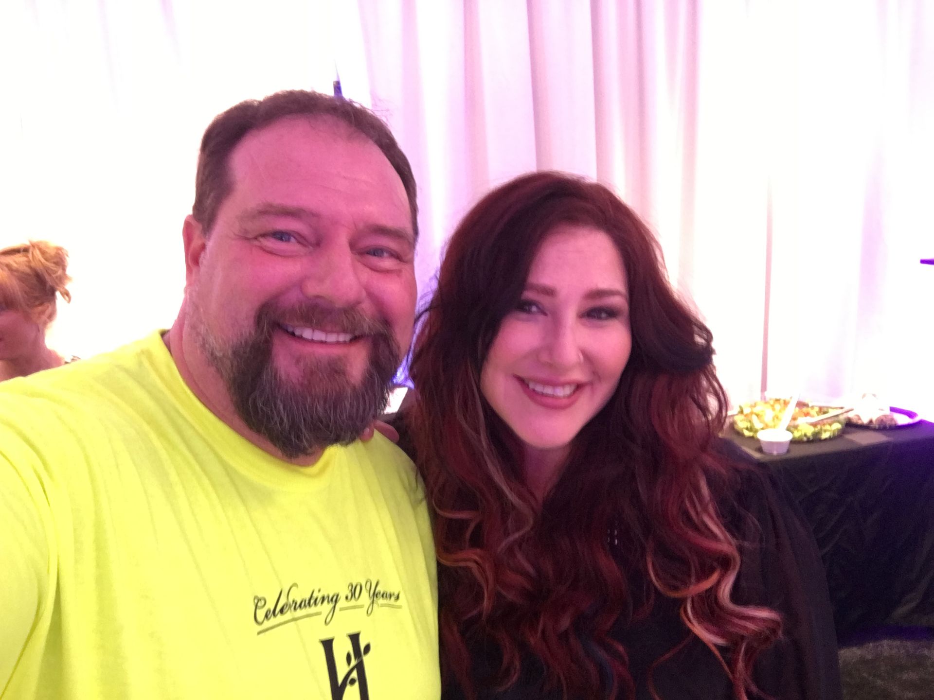 A man in a bright yellow T-shirt and a woman with long red-brown hair smiling together indoors in front of white drapes.