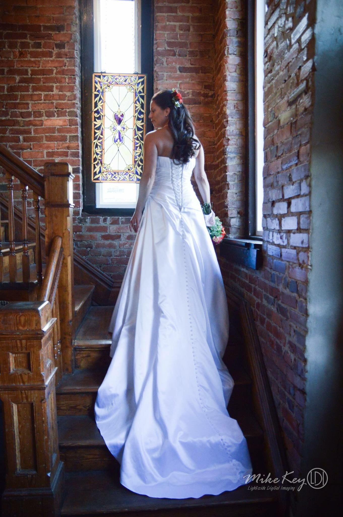 A person in a white wedding gown stands on wooden stairs between brick walls, looking toward a stained-glass window.