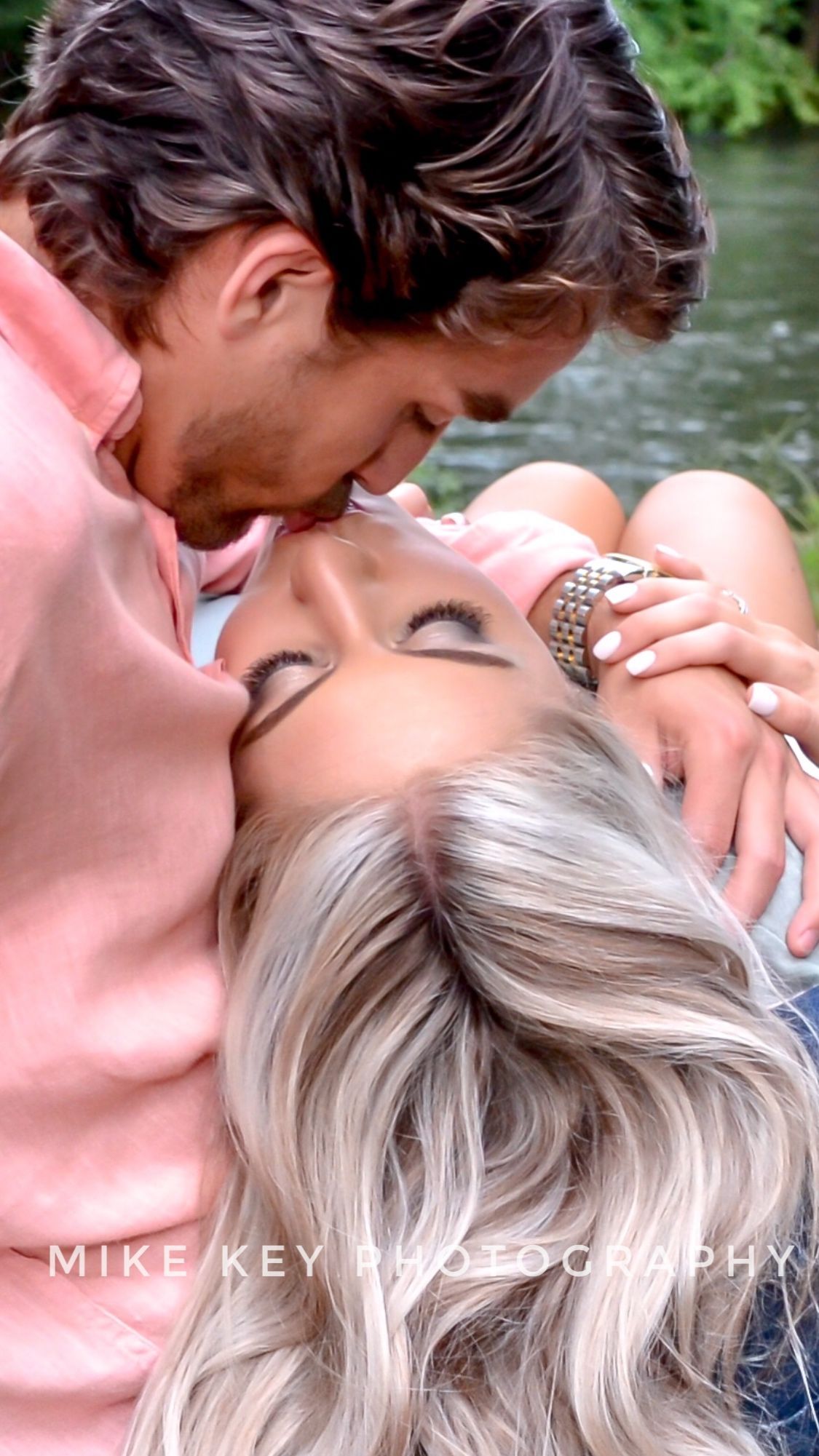 A man in a pink shirt kisses a woman with blonde hair, who is lying in his arms outdoors near water.