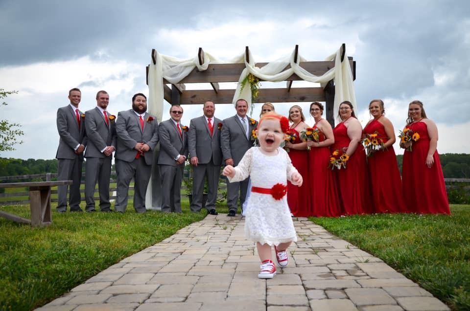 A toddler runs toward the camera on a stone path in front of a wedding party standing under an outdoor wooden arbor.