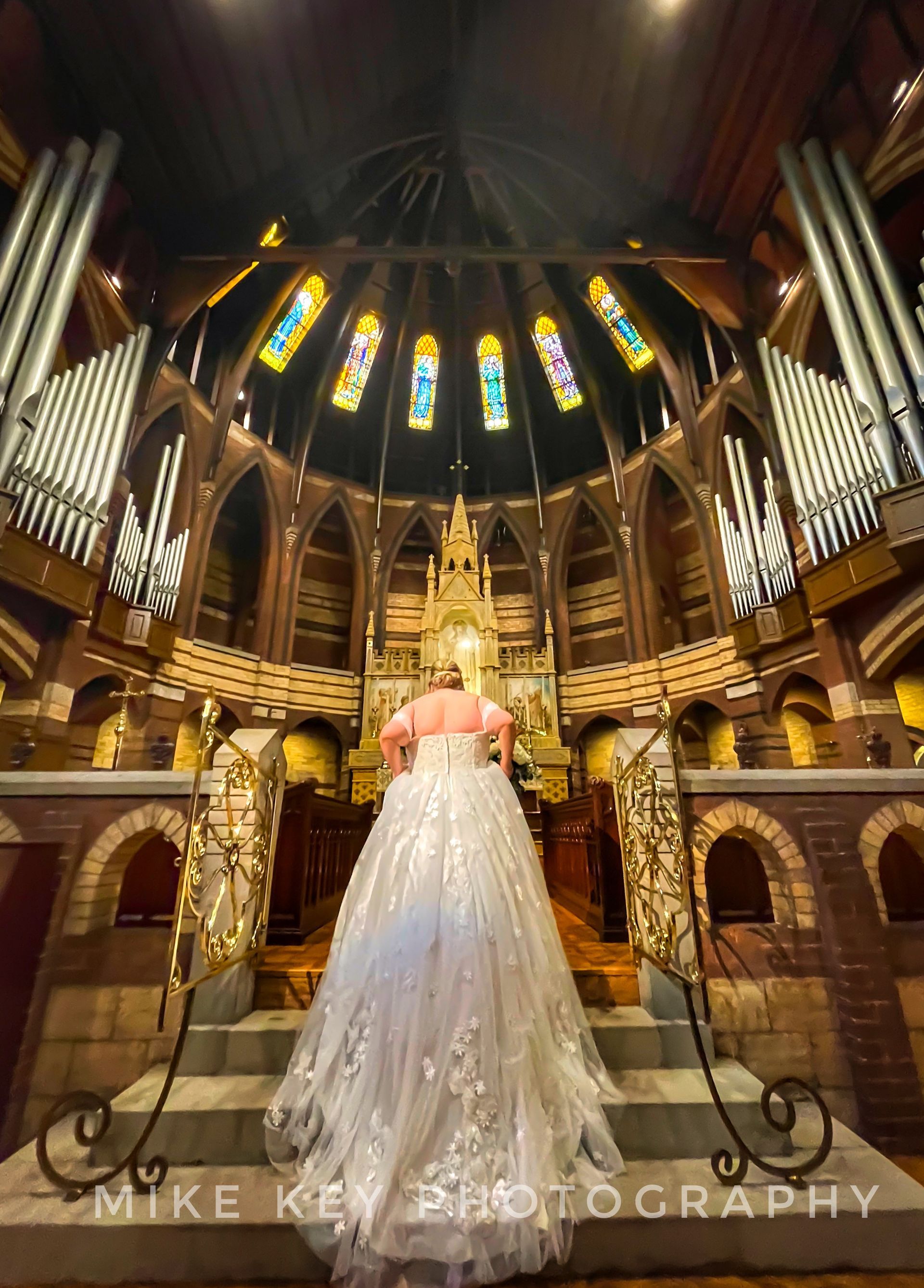 A person in a large, white wedding dress stands at the top of stone steps inside a majestic church with pipe organs.