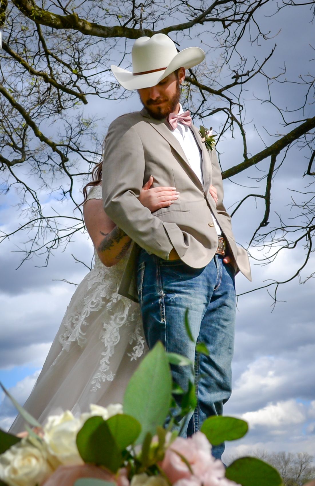 A bride in a white lace dress stands behind a man in a tan blazer, cowboy hat, and jeans, captured against a cloudy sky.