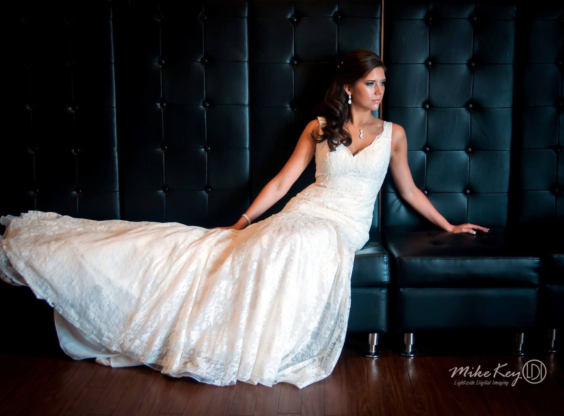 A person in a white lace bridal gown reclines on a tufted black leather bench against a dark paneled wall.