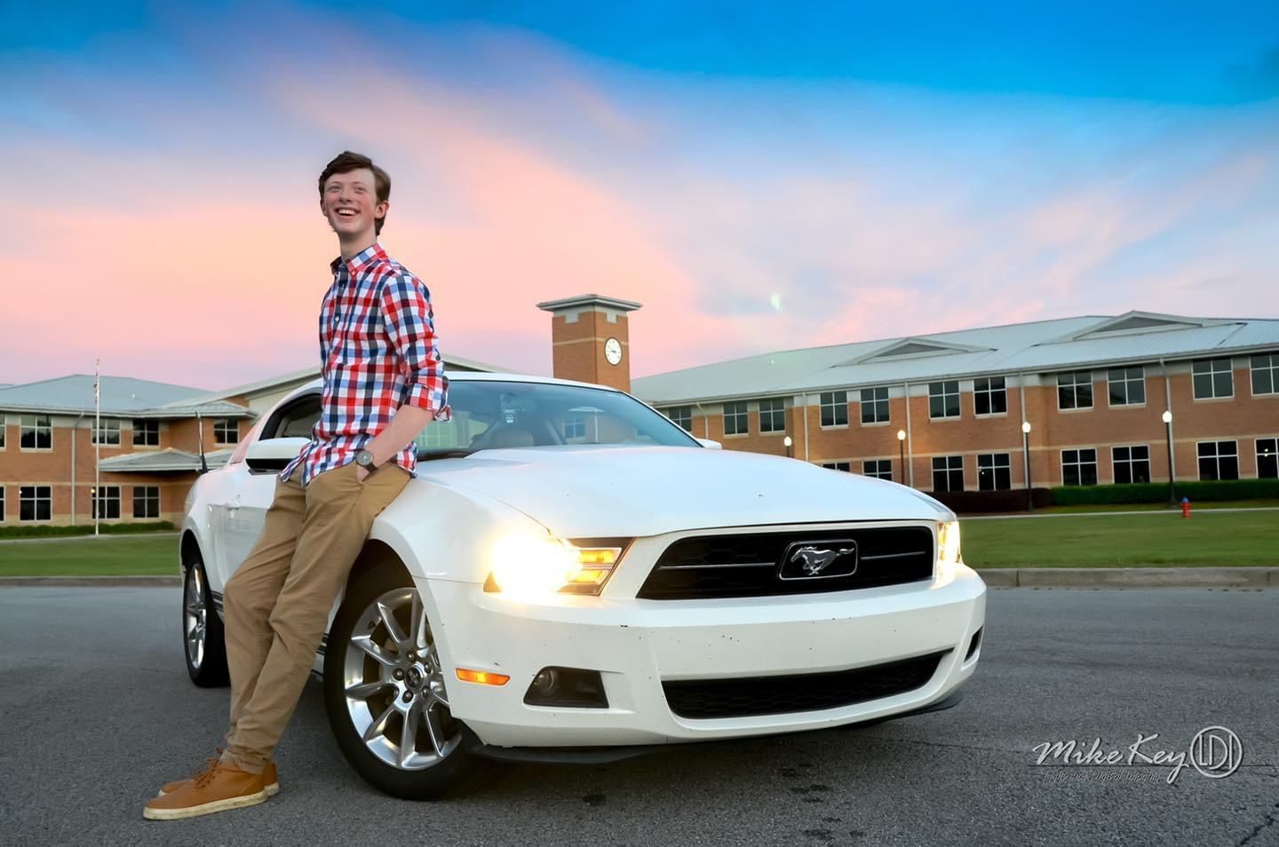 A person in a red plaid shirt and khaki pants leans against the hood of a white Ford Mustang at sunset.
