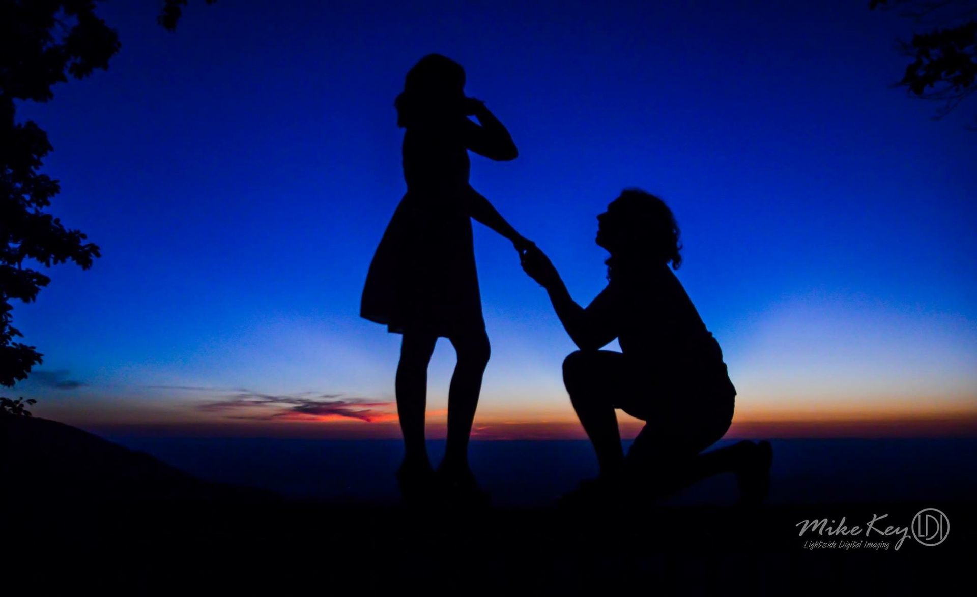 A silhouette of one person kneeling on one knee, holding the hand of another person standing against a sunset sky.