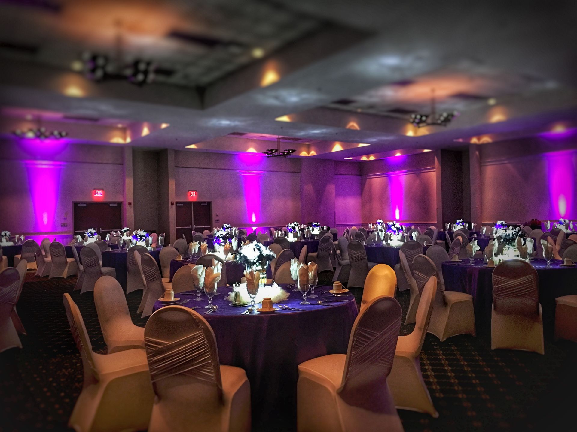An event hall set for a banquet with round tables, purple tablecloths, and chairs with gold covers, bathed in pink light.