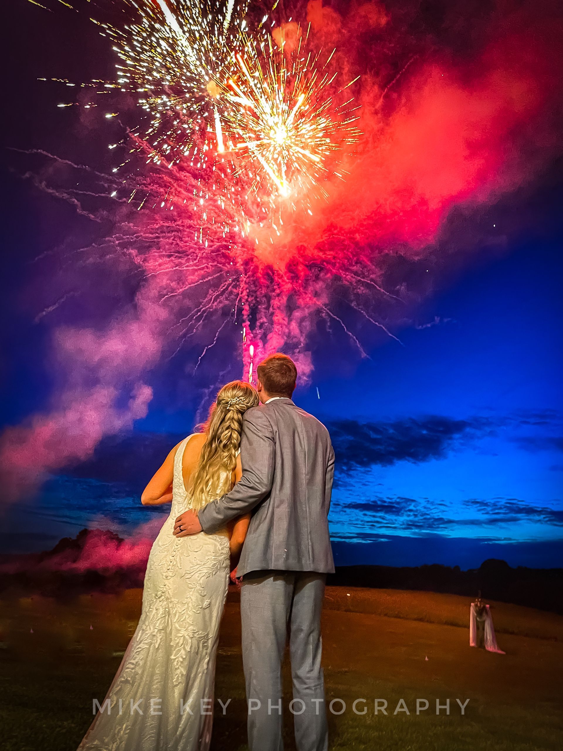 A couple stands outdoors at dusk, looking up at a bright, vibrant explosion of fireworks in a deep blue night sky.