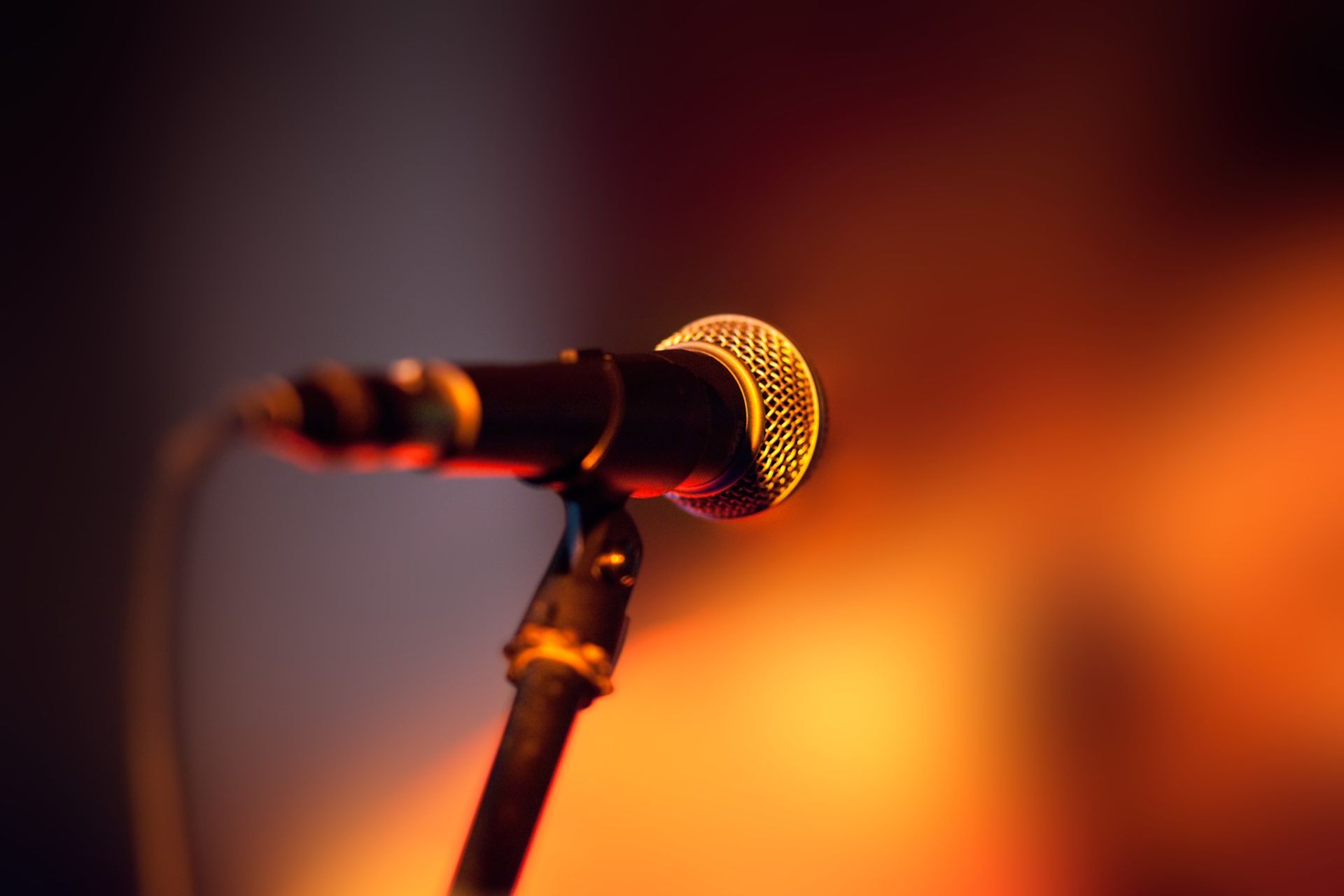 A close-up view of a stage microphone on a stand, illuminated by warm orange lighting against a dark, blurred background.