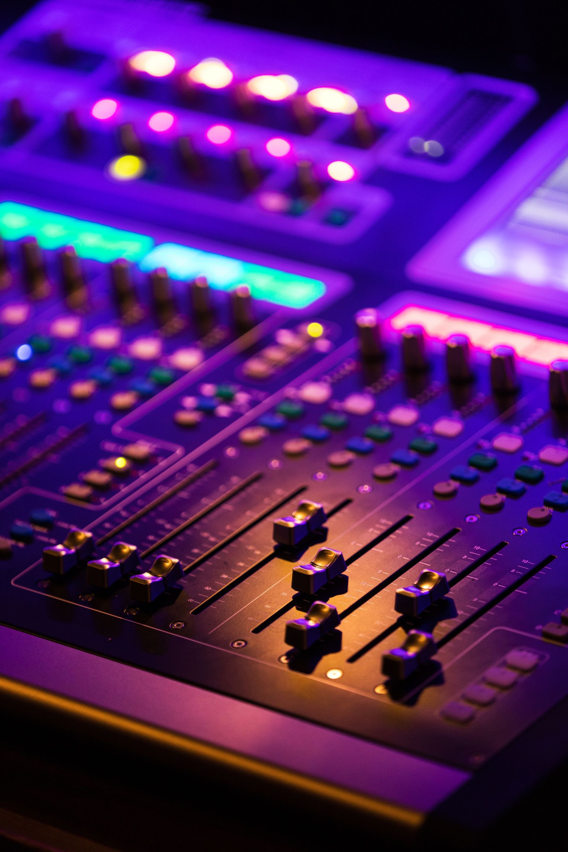 A close-up of a professional audio mixing console with illuminated sliders and knobs bathed in purple and amber light.