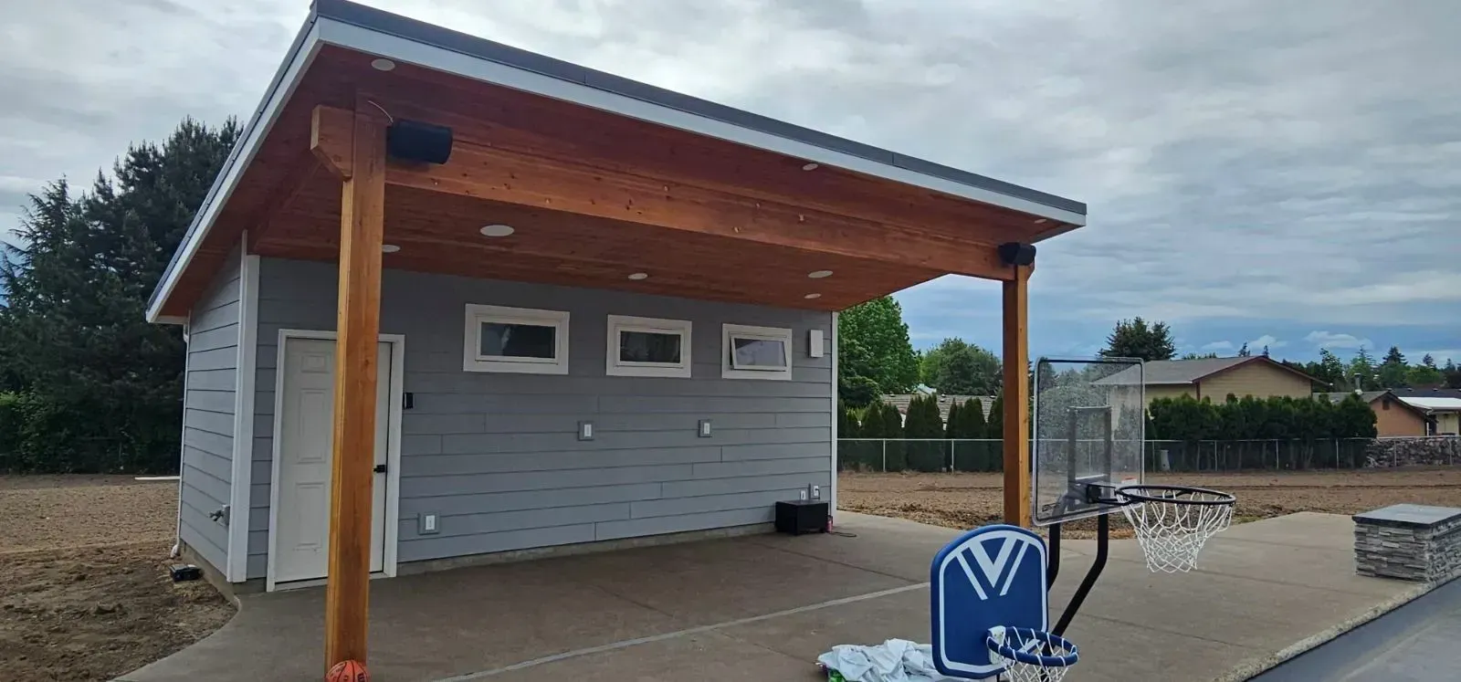 A gray outdoor pavilion with a wooden roof, a single white door, three small windows, and a basketball hoop on a patio.
