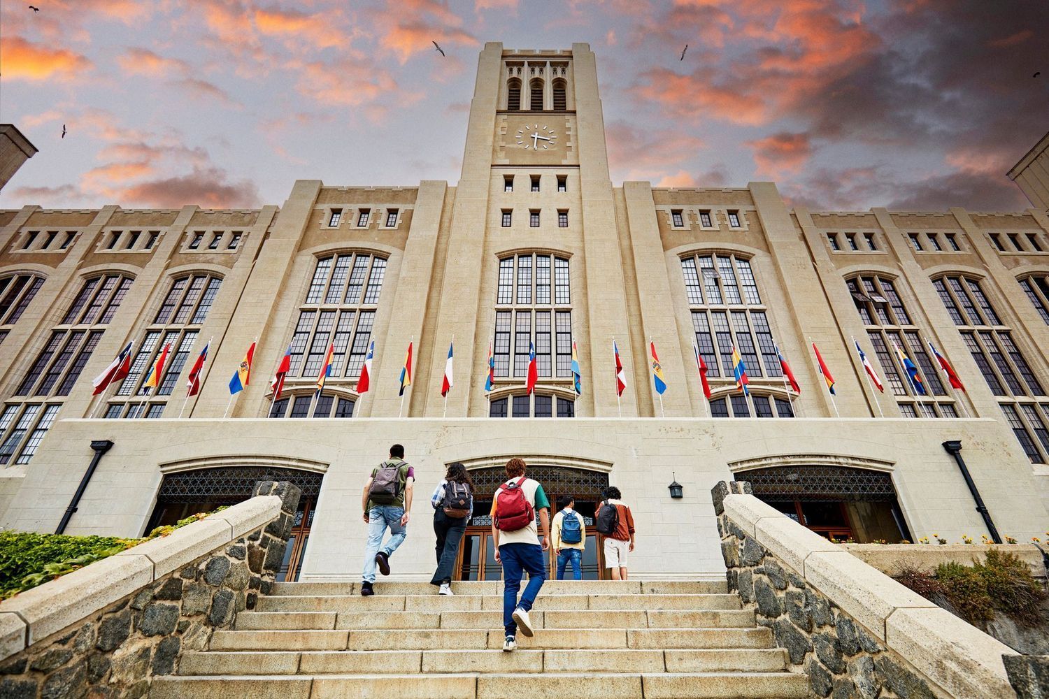 Students walk up the stone steps of a large university building with rows of international flags displayed in front.