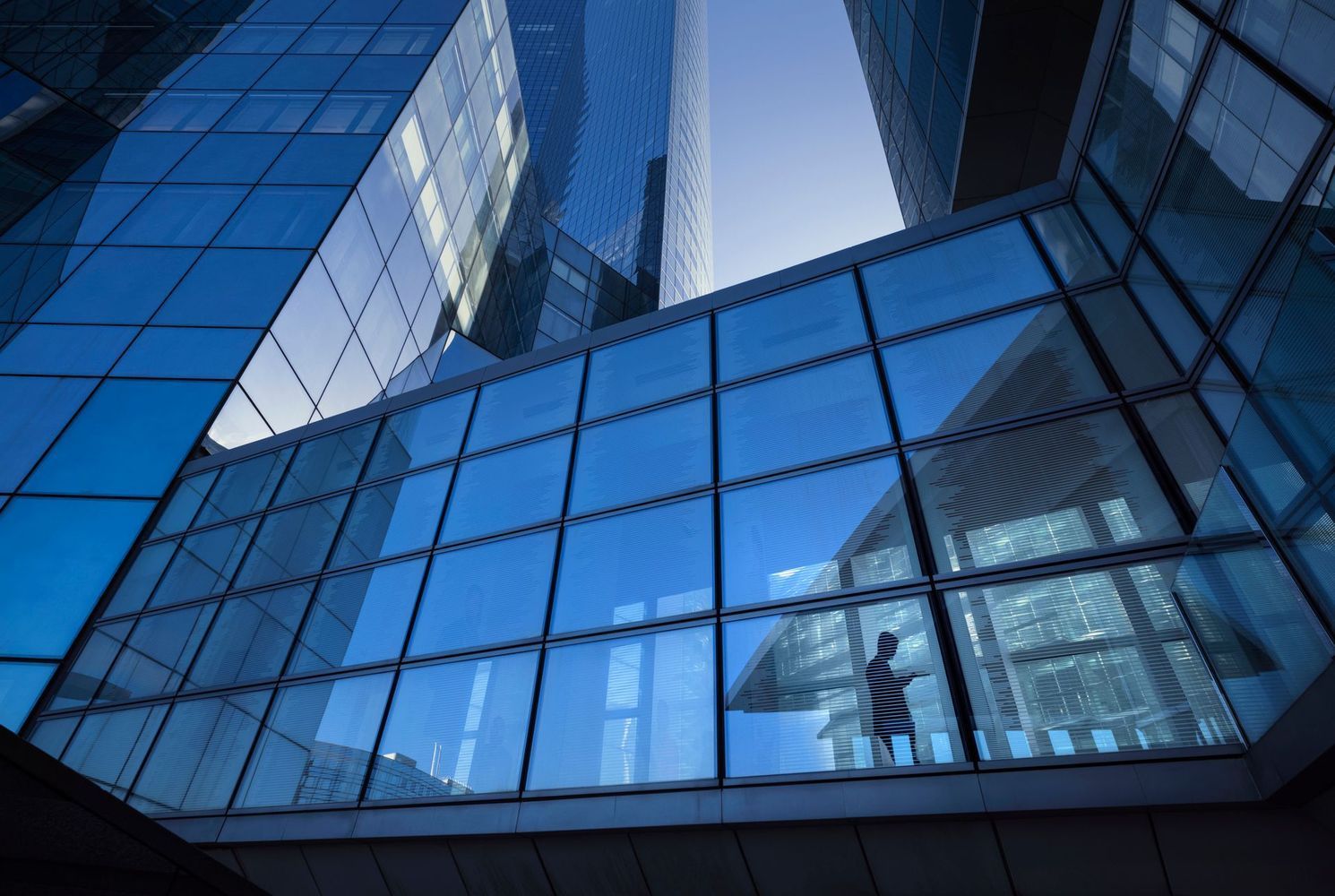 A lone person walks across a glass skybridge connecting two tall, modern blue glass skyscrapers against a clear sky.