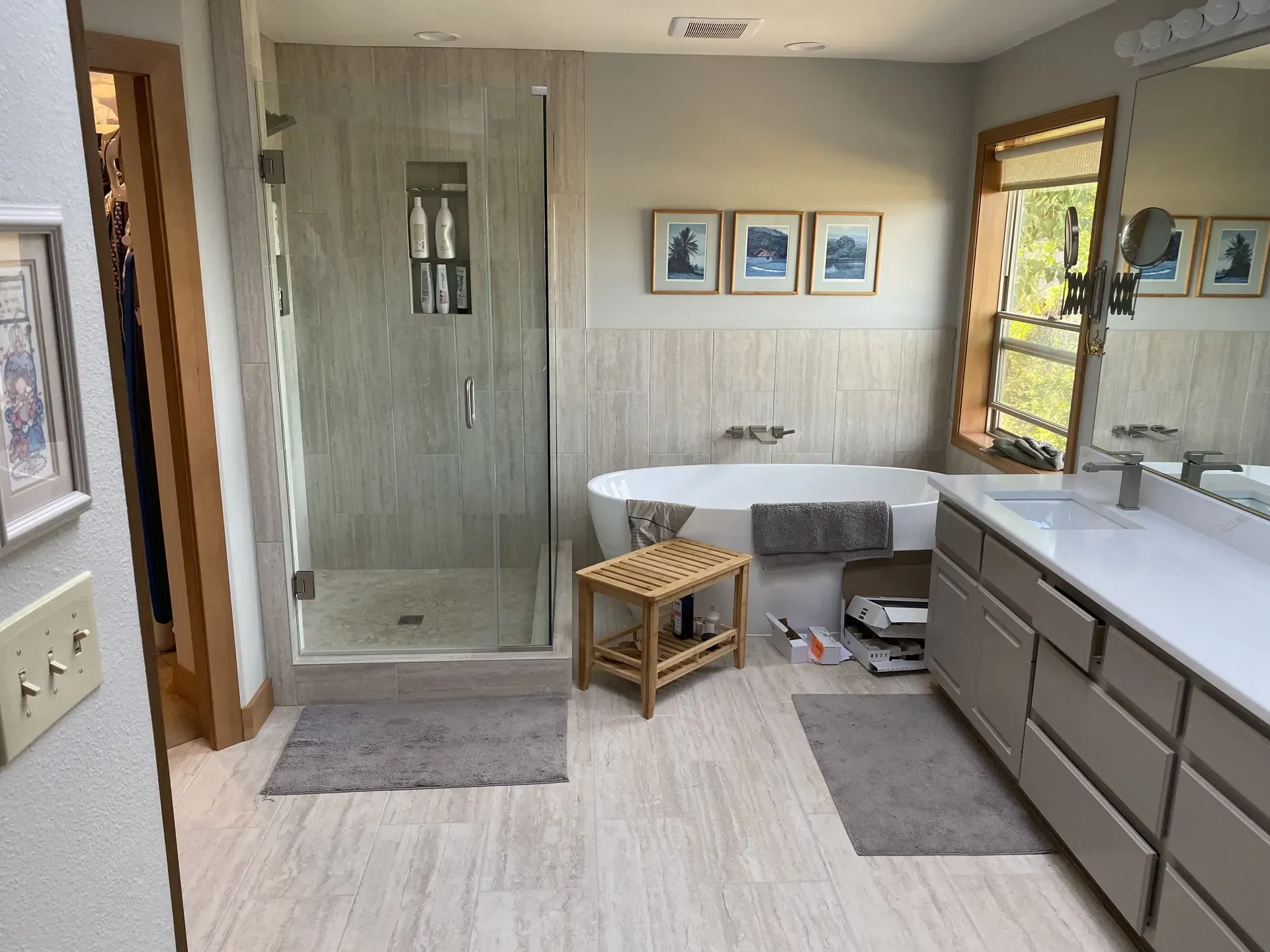 Modern bathroom featuring a glass shower, soaking tub, wooden stool, vanity with double sinks, and light-toned tile floor.