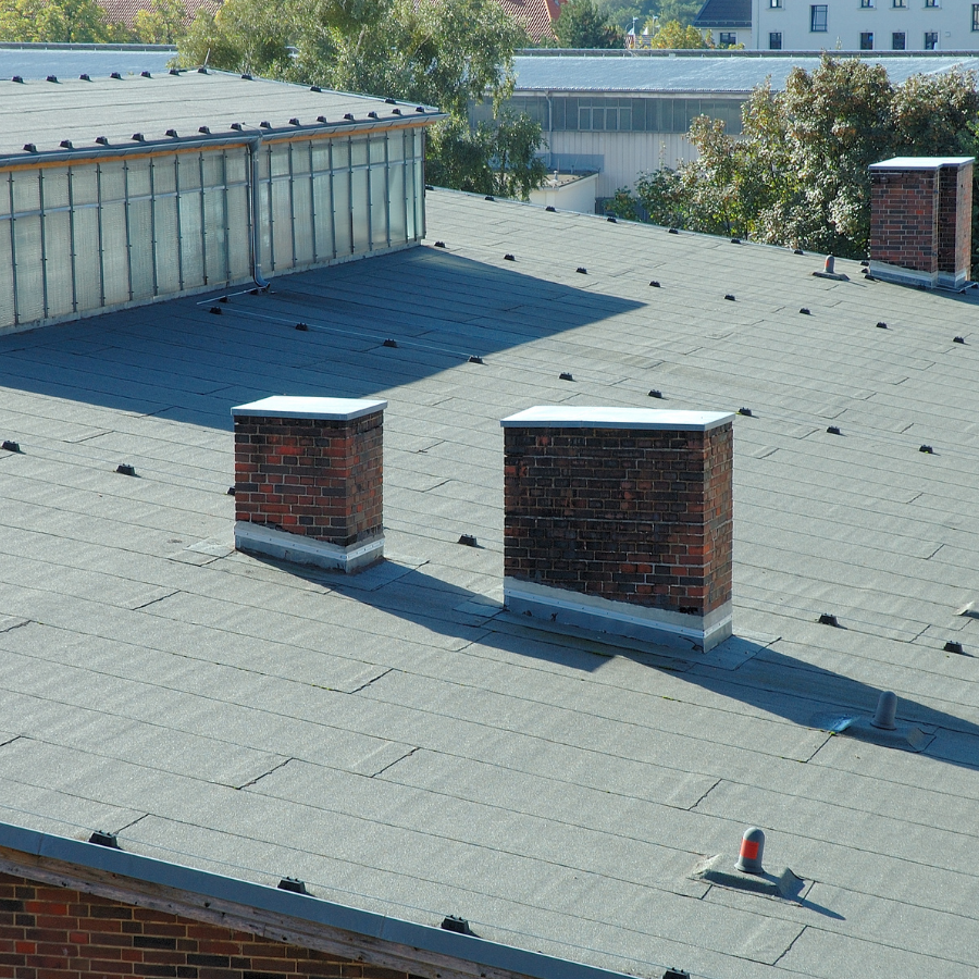 Flat rooftop with gray shingles, two brick chimneys, and a glass skylight under clear daylight
