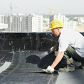 Worker in a yellow hard hat applying roofing material on a rooftop, with city buildings in the background