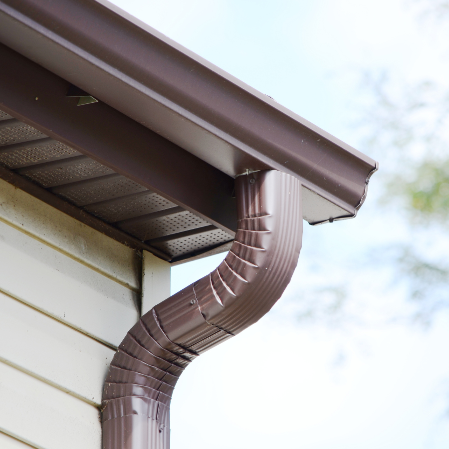 Brown rain gutter and downspout on a house roof corner against a cloudy sky