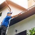 Worker in blue shirt cleaning roof gutters from a ladder beside a house