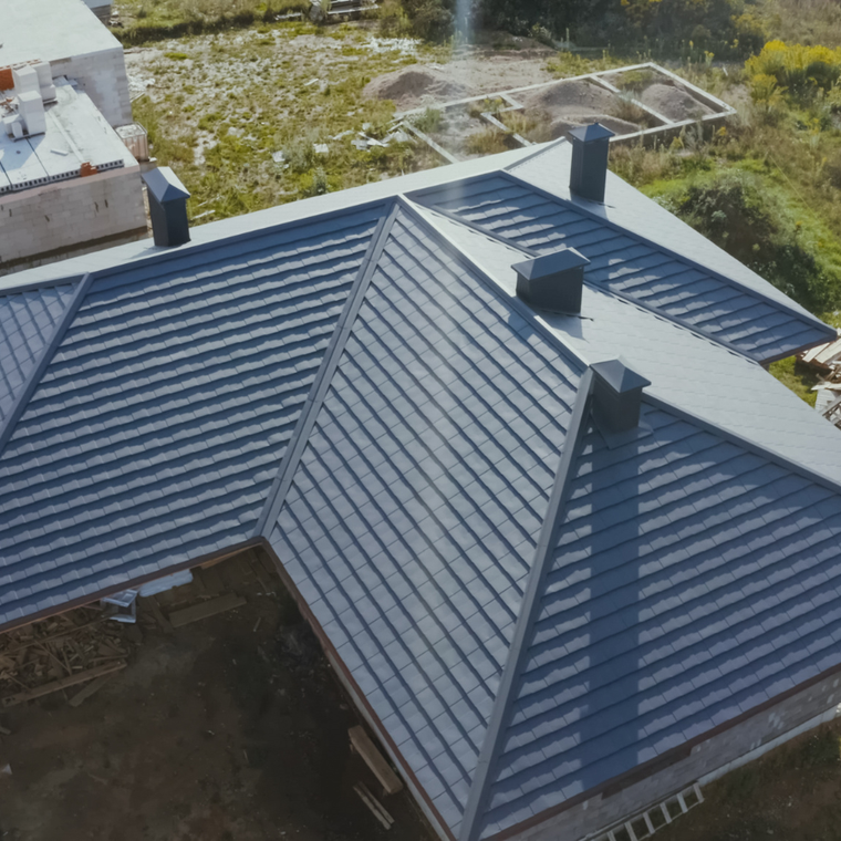 Aerial view of a dark metal roof with three chimneys and a small skylight, beside grassy yard and garden beds