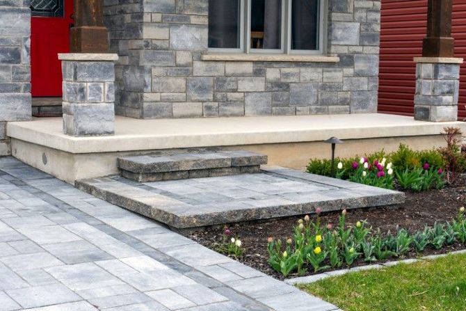 A stone-veneer porch with a concrete platform, paver walkway, and stone step, flanked by flower beds and green lawn.