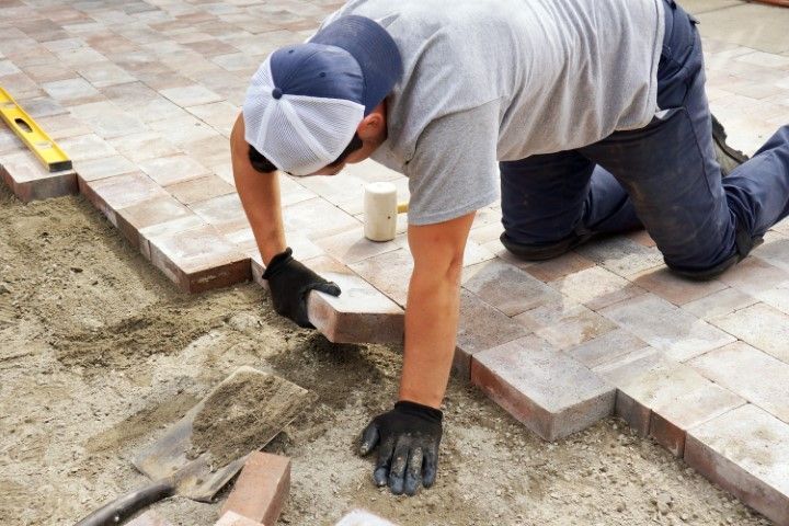 A person in a gray shirt, hat, and work gloves kneeling on the ground while placing rectangular pavers into a sand bed.