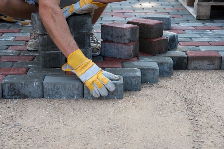A person wearing safety gloves installs gray rectangular paving stones on a leveled gravel base.