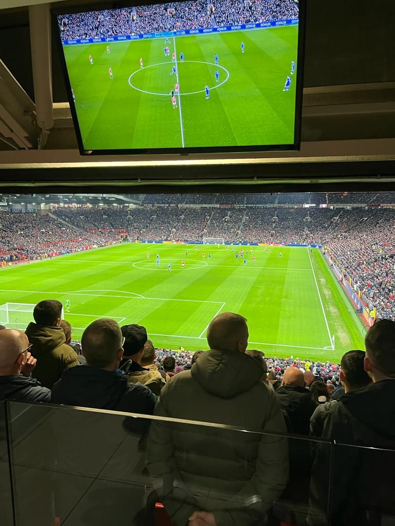 Crowd watching a soccer match from stadium seats, with the field and players visible below and on a screen above.