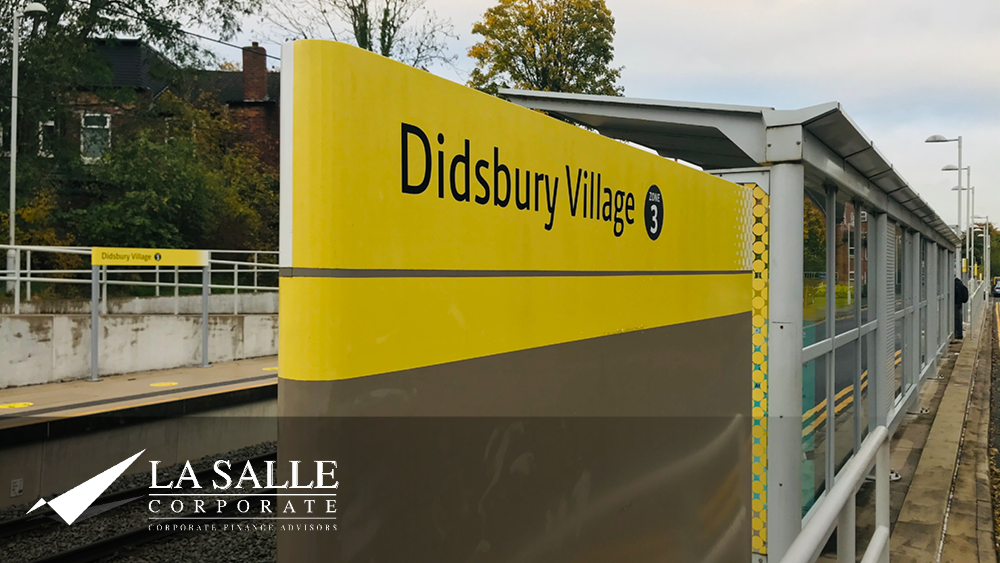 Didsbury Village station sign on a yellow platform shelter beside train tracks