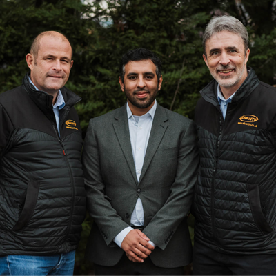 Three men posing outdoors, two in black jackets and one in a gray suit, in front of trees