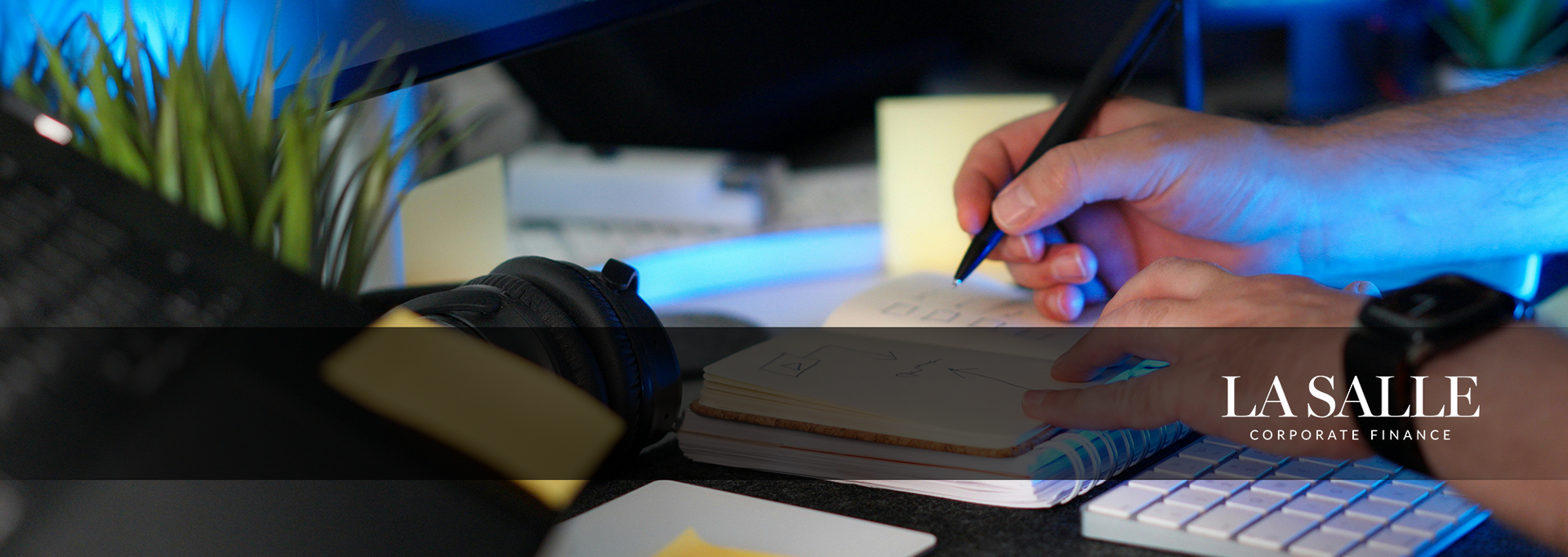 Hands writing in a notebook beside a laptop on a desk with a blue blurred background.