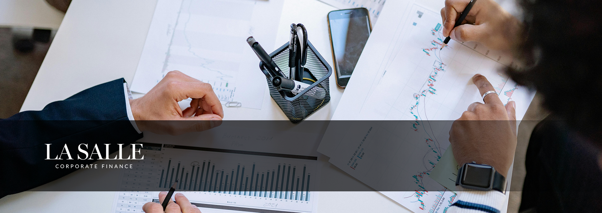 Hands reviewing financial charts with a calculator and pen on a desk, with a laptop and business logo overlay.
