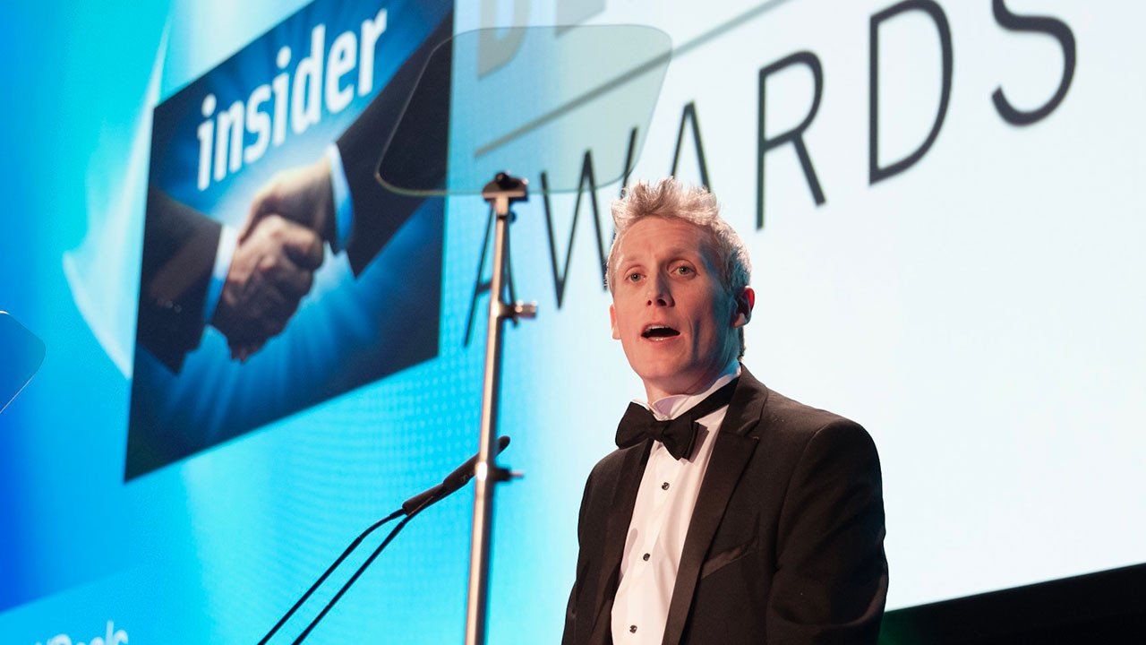 Man in tuxedo speaks at podium before a “TOWARDS” event banner and microphone