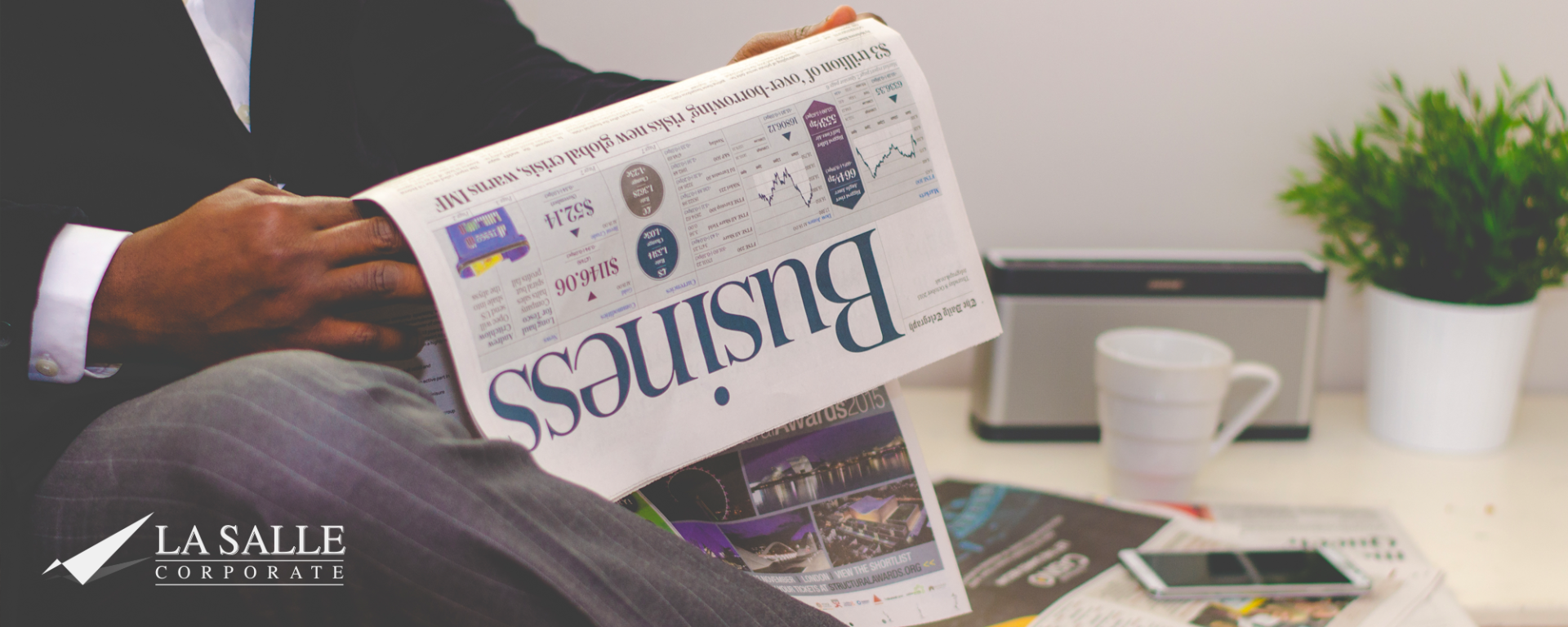 Person reading a Business newspaper at a desk with a laptop, coffee cup, and potted plant.