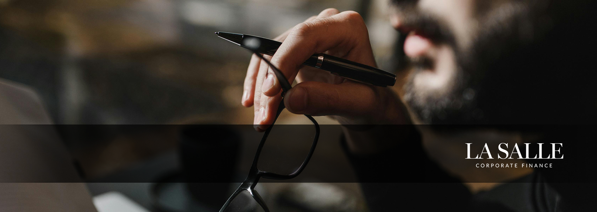 Hand holding a black pen over eyeglasses, with a blurred dark background and logo text on the right.